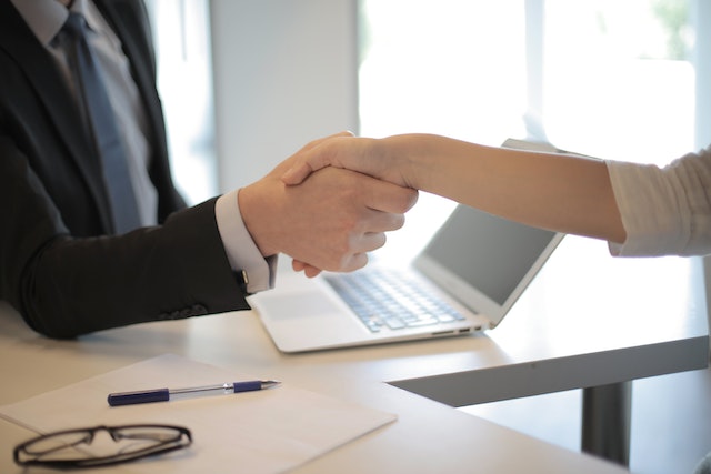 a property manager in a black suit reaches across the contract and pen laying on their desk to shake a landlords hand